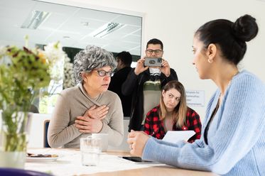 Eine angehende Pflegerin übt ein Gespräch mit einer Schauspielerin, die als alte Frau verkleidet ist. Mitschüler im Hintergrund filmen die Szene und machen Notizen.